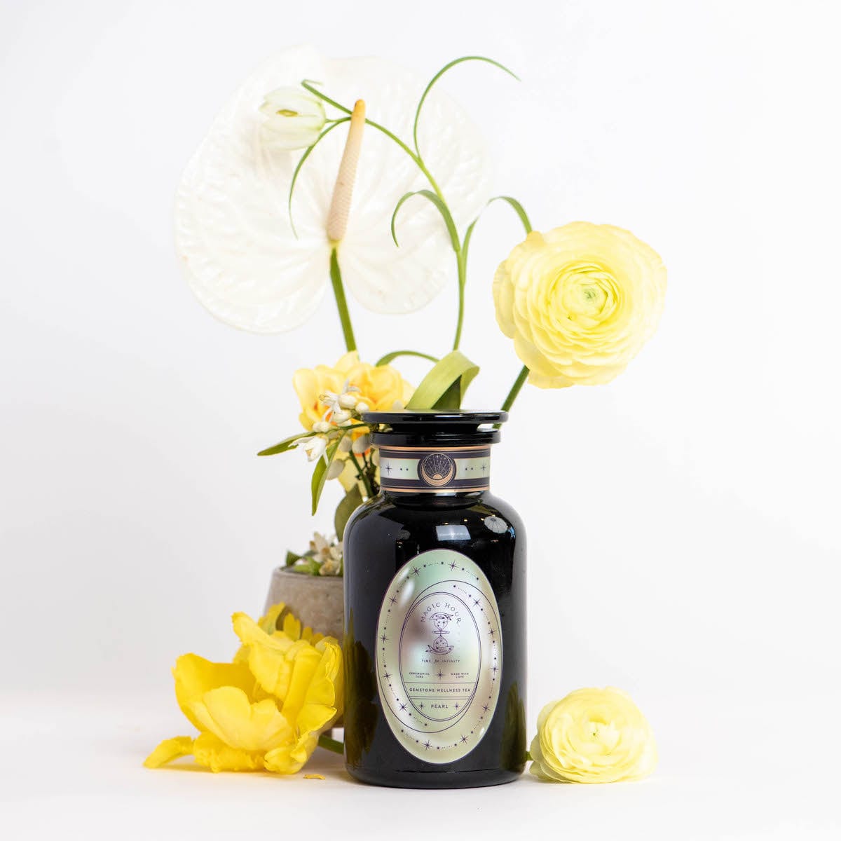 A black jar labeled "Pearl Tea of Beauty, Clarity, and a Sense of Peace" from Magic Hour stands on a white surface, surrounded by various yellow and white flowers, including ranunculus and calla lilies. The flowers complement the elegant jar, creating a visually appealing arrangement against a plain white background.