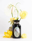 A black jar labeled "Pearl Tea of Beauty, Clarity, and a Sense of Peace" from Magic Hour stands on a white surface, surrounded by various yellow and white flowers, including ranunculus and calla lilies. The flowers complement the elegant jar, creating a visually appealing arrangement against a plain white background.