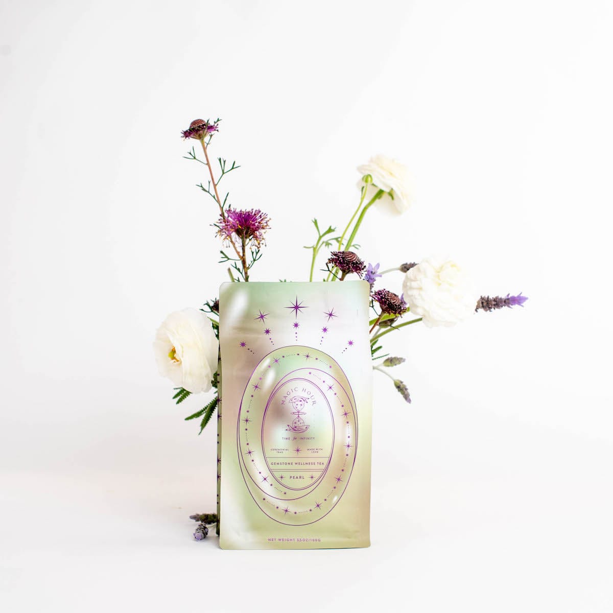 A green foil packet labeled "Magic Hour Pearl Tea of Beauty, Clarity, and a Sense of Peace" is displayed in front of a minimalistic floral arrangement with white, purple, and lavender flowers, all set against a plain white background, evoking the calm elegance reminiscent of a Magic Hour Tea ceremony.