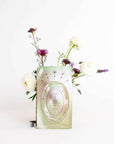 A green foil packet labeled "Magic Hour Pearl Tea of Beauty, Clarity, and a Sense of Peace" is displayed in front of a minimalistic floral arrangement with white, purple, and lavender flowers, all set against a plain white background, evoking the calm elegance reminiscent of a Magic Hour Tea ceremony.