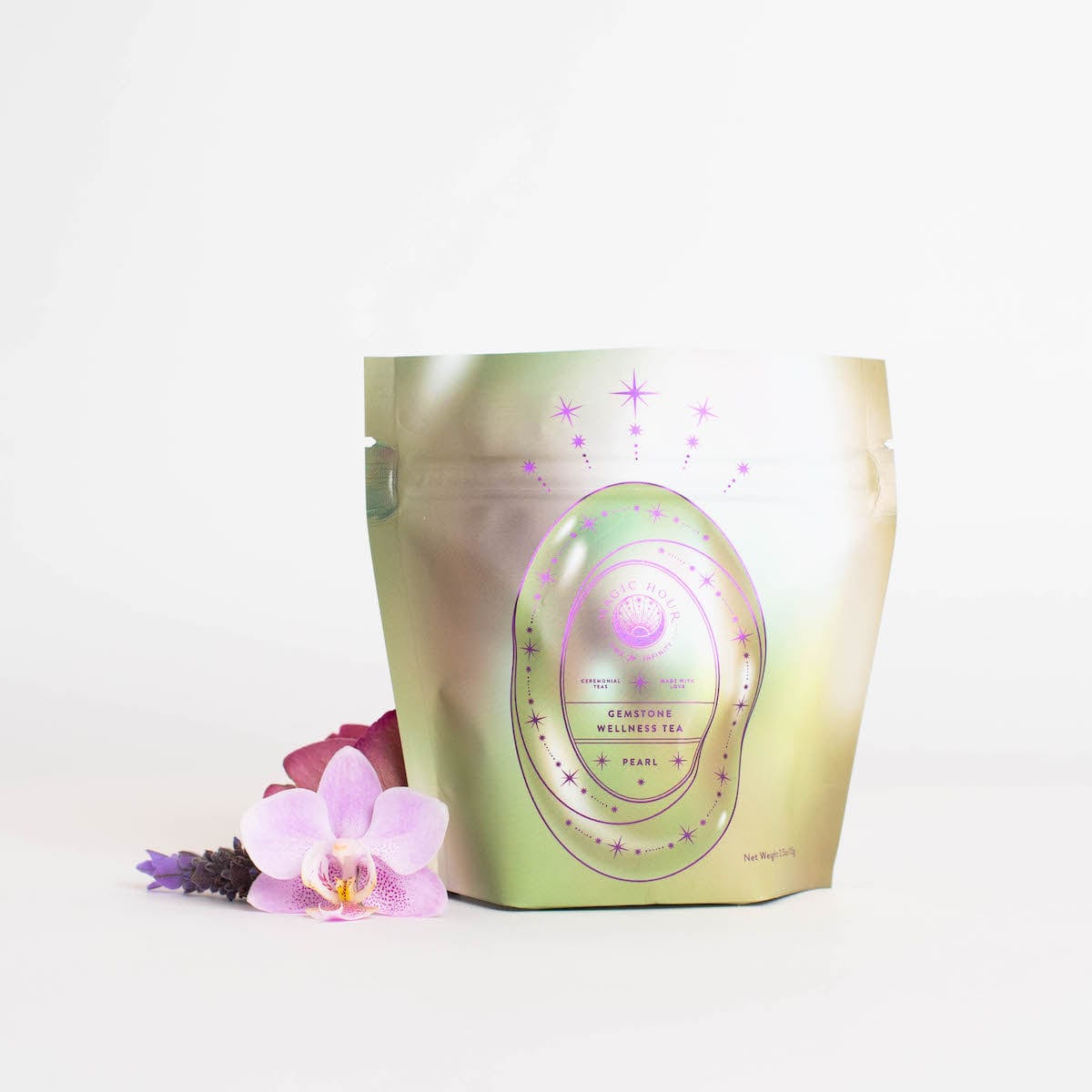 A shiny, metallic pouch of "Magic Hour Pearl Tea of Beauty, Clarity, and a Sense of Peace" is displayed against a white background. The loose leaf organic tea pouch features a pink and purple design with stars and an oval-shaped label. Beside it lie pink orchids and a purple flower, adding a touch of elegance.