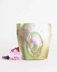 A shiny, metallic pouch of "Magic Hour Pearl Tea of Beauty, Clarity, and a Sense of Peace" is displayed against a white background. The loose leaf organic tea pouch features a pink and purple design with stars and an oval-shaped label. Beside it lie pink orchids and a purple flower, adding a touch of elegance.