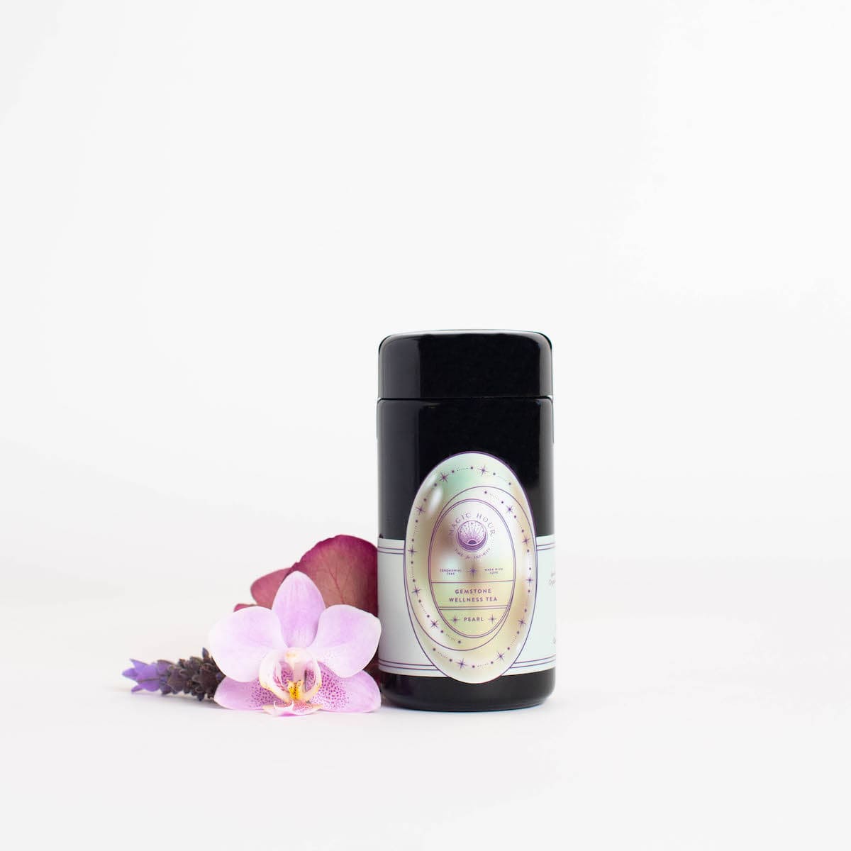 A sleek, black cylindrical product container with an elegant label featuring a circular, multi-colored design stands upright. Next to it, there are a purple orchid flower, a lavender sprig, and a red rose petal from Magic Hour's Pearl Tea of Beauty, Clarity, and a Sense of Peace, all on a white background.