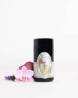 A sleek, black cylindrical product container with an elegant label featuring a circular, multi-colored design stands upright. Next to it, there are a purple orchid flower, a lavender sprig, and a red rose petal from Magic Hour's Pearl Tea of Beauty, Clarity, and a Sense of Peace, all on a white background.