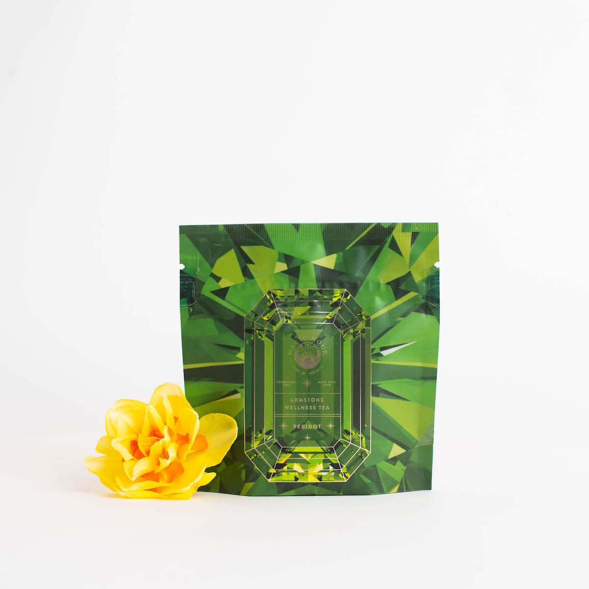 A green, gem-shaped patterned tea package from Magic Hour's Peridot: Green Guayusa Oolong Mango-Peach Tea Case Packs stands upright against a white background. To the left of the package is a bright yellow flower.