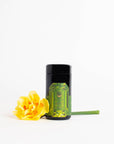 A refined black cylindrical container with a striking green label featuring the Magic Hour logo stands upright on a white background. To its left, a vibrant yellow flower with a green stem rests horizontally, infusing the minimalist scene with color and nature. The label offers a tantalizing hint of the refreshing aroma of organic mango within, announcing it as Peridot: Green Guayusa Oolong Mango-Peach Tea Case Packs.