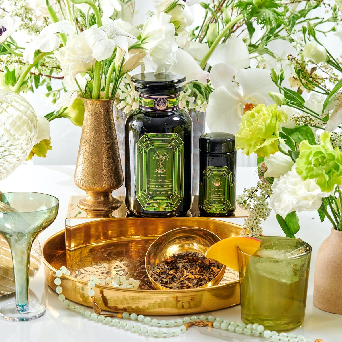 A lavish tabletop display featuring a brass tray holding a matching spherical container of Magic Hour's Peridot: Green Guayusa Oolong Mango-Peach Tea case packs, a golden goblet, and a green and black jar. Surrounding the tray are white and green flowers in vases, an organic mango cocktail, a beaded necklace, and a green wine glass.
