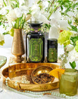 A lavish tabletop display featuring a brass tray holding a matching spherical container of Magic Hour's Peridot: Green Guayusa Oolong Mango-Peach Tea case packs, a golden goblet, and a green and black jar. Surrounding the tray are white and green flowers in vases, an organic mango cocktail, a beaded necklace, and a green wine glass.