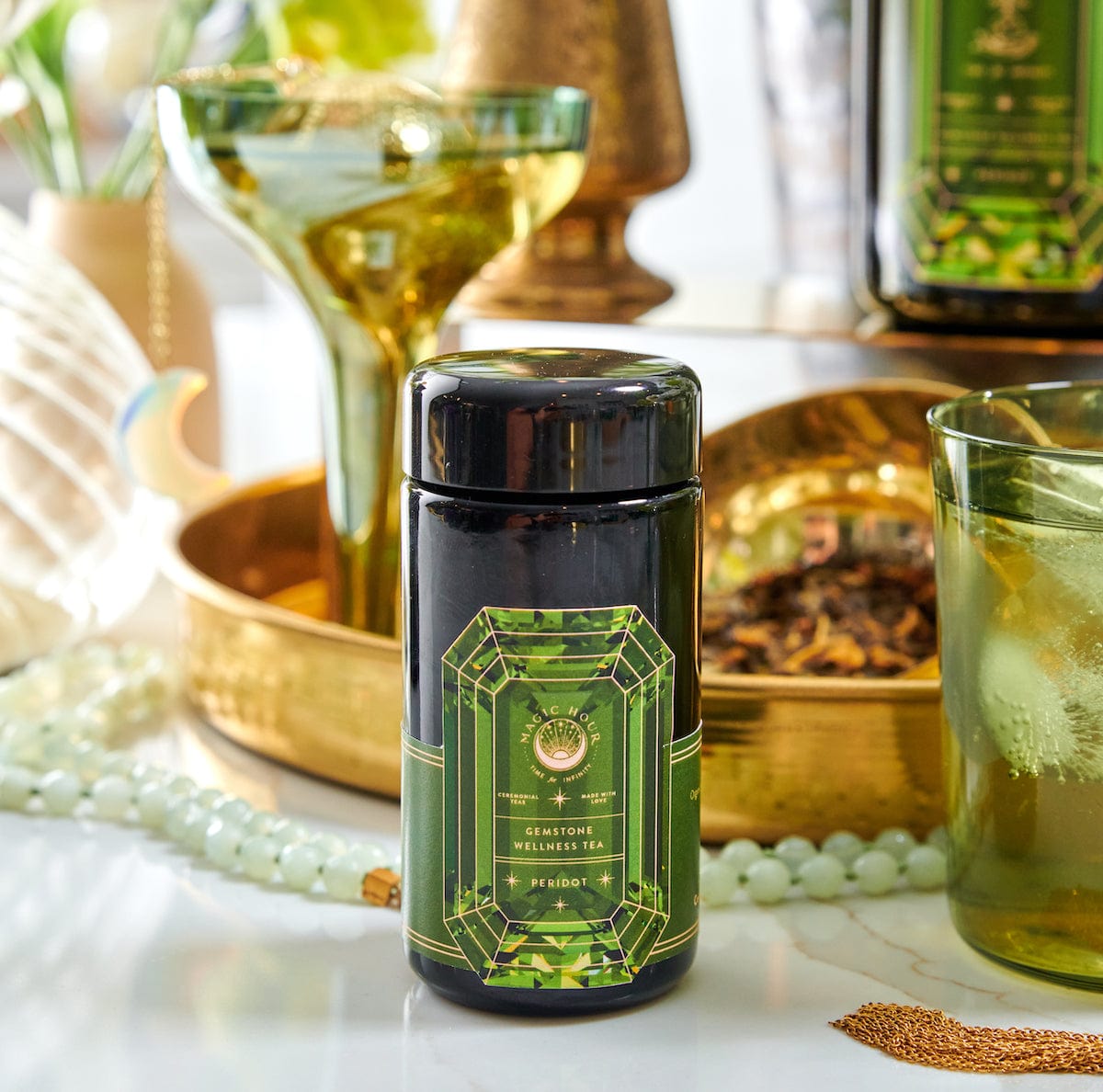 A black jar of Magic Hour's Peridot: Green Guayusa Oolong Mango-Peach Tea is surrounded by various green and gold decorations, including beads, a glass of Organic Mango tea, and a green bottle. The jar's label features an ornate green gemstone design and detailed text.