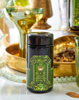 A black jar of Magic Hour's Peridot: Green Guayusa Oolong Mango-Peach Tea is surrounded by various green and gold decorations, including beads, a glass of Organic Mango tea, and a green bottle. The jar's label features an ornate green gemstone design and detailed text.