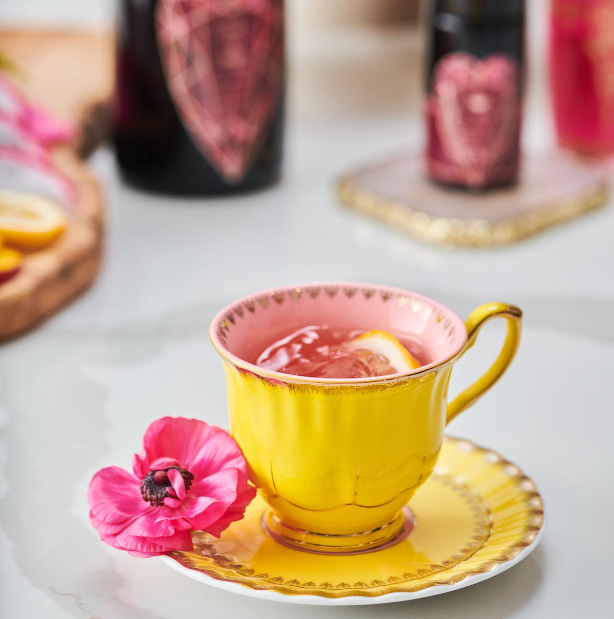 A yellow teacup with a pink interior sits on a matching saucer, filled with Pink Tourmaline: Organic Dragon Fruit Hibiscus White Tea Case Packs by Magic Hour and garnished with a lemon slice. A pink flower is placed beside the teacup on the saucer. Blurred bottles and a cutting board with lemon slices are visible in the background.