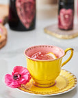 A yellow teacup with a pink interior sits on a matching saucer, filled with Pink Tourmaline: Organic Dragon Fruit Hibiscus White Tea Case Packs by Magic Hour and garnished with a lemon slice. A pink flower is placed beside the teacup on the saucer. Blurred bottles and a cutting board with lemon slices are visible in the background.