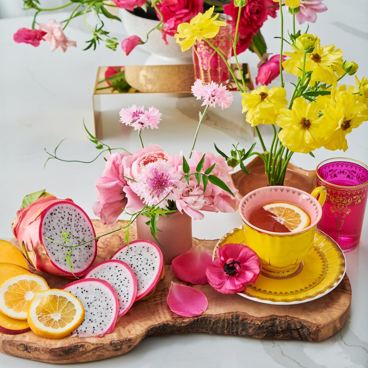 A vibrant tabletop setup features a yellow teacup with an immune-supportive blend of Magic Hour's Pink Tourmaline: Organic Dragon Fruit Hibiscus White Tea, surrounded by fresh flowers in yellow and pink. Slices of dragon fruit, mango, and lemon are artfully arranged on a wooden board. A pink glass adds an additional pop of color to this bright and cheerful arrangement.