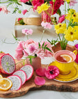 A vibrant tabletop setup features a yellow teacup with an immune-supportive blend of Magic Hour's Pink Tourmaline: Organic Dragon Fruit Hibiscus White Tea, surrounded by fresh flowers in yellow and pink. Slices of dragon fruit, mango, and lemon are artfully arranged on a wooden board. A pink glass adds an additional pop of color to this bright and cheerful arrangement.
