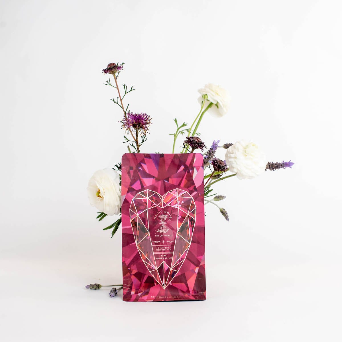 A pink geometric gift bag with a heart design stands against a white background. The bag is flanked by a simple flower arrangement with white and purple blooms and greenery, creating a visually pleasing contrast reminiscent of the soothing properties found in Magic Hour's Pink Tourmaline: Organic Dragon Fruit Hibiscus White Tea Case Packs.
