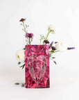 A pink geometric gift bag with a heart design stands against a white background. The bag is flanked by a simple flower arrangement with white and purple blooms and greenery, creating a visually pleasing contrast reminiscent of the soothing properties found in Magic Hour's Pink Tourmaline: Organic Dragon Fruit Hibiscus White Tea Case Packs.