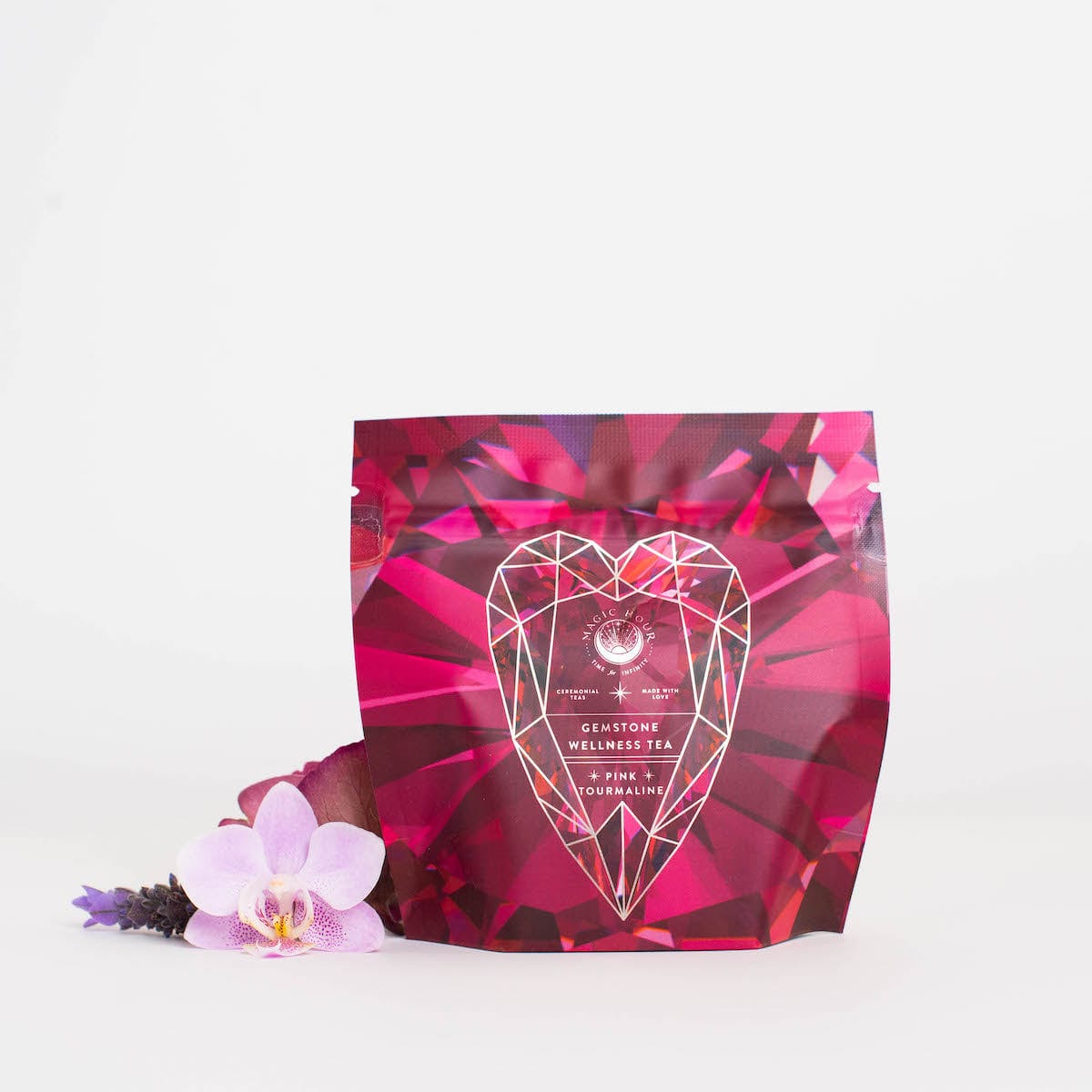 A package of Magic Hour's Pink Tourmaline: Organic Dragon Fruit Hibiscus White Tea Case Packs, known for their heart and breast health benefits, is pictured. The packaging features a predominantly pink design with a faceted gemstone graphic on the front. An orchid flower and a small sprig of lavender are placed beside the package against a white background.