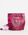 A package of Magic Hour's Pink Tourmaline: Organic Dragon Fruit Hibiscus White Tea Case Packs, known for their heart and breast health benefits, is pictured. The packaging features a predominantly pink design with a faceted gemstone graphic on the front. An orchid flower and a small sprig of lavender are placed beside the package against a white background.