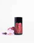 A black cylindrical container from the Magic Hour brand, adorned with a red label featuring a geometric heart design, stands beside three flowers: a white and pink orchid, a purple flower, and a red flower. The background is plain white. This Pink Tourmaline: Organic Dragon Fruit Hibiscus White Tea Case Pack promotes Heart & Breast Health while adding an elegant touch to your decor.