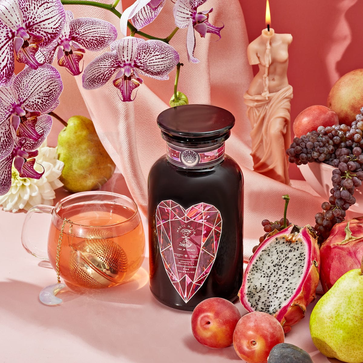 A still-life composition showcasing a jar with a heart-shaped label, symbolizing Heart & Breast Health, surrounded by orchids, a lit candle, a cup of Magic Hour's Pink Tourmaline: Organic Dragon Fruit Hibiscus White Tea Case Packs, and an assortment of fresh fruits including a pear, dragon fruit, grapes, and plums—all set against a pink background.