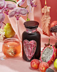 A still-life composition showcasing a jar with a heart-shaped label, symbolizing Heart & Breast Health, surrounded by orchids, a lit candle, a cup of Magic Hour's Pink Tourmaline: Organic Dragon Fruit Hibiscus White Tea Case Packs, and an assortment of fresh fruits including a pear, dragon fruit, grapes, and plums—all set against a pink background.