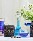 A collection of elegant glassware with blue liquids, including a pitcher, decanter, glasses, and bottles is arranged on a white surface with a bouquet of white flowers in the background. A packet of "Pisces Tea - Oolong, Magnolia and Jasmine Sampler Pouch" is also shown.
