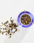 Overhead view of a glass teacup with blue tea brewed from The Astrology Collection - Luxe Sampler Sips, featuring an elegant metal infuser holding rare teas and petals. Scattered on the white surface beside the cup are some loose tea leaves, adding to the visual delight.