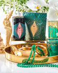 A golden tray displays an assortment of ornate items, including a green and gold teacup, a green beaded necklace, a gold teapot with Organic Ceremonial Matcha 700 from Club Magic Hour, and a jar with a teal and pink label. Floral arrangements with white flowers and statues are in the background.