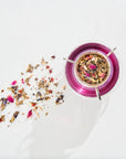 A clear glass teapot filled with hot pink herbal tea sits on a white surface. Next to the teapot, Magic Hour's Luxe Tea Sampler Sips: The Queens Collection colorful loose leaf tea ingredients, including dried petals and herbs, are scattered, adding a vibrant touch to the minimalist background.