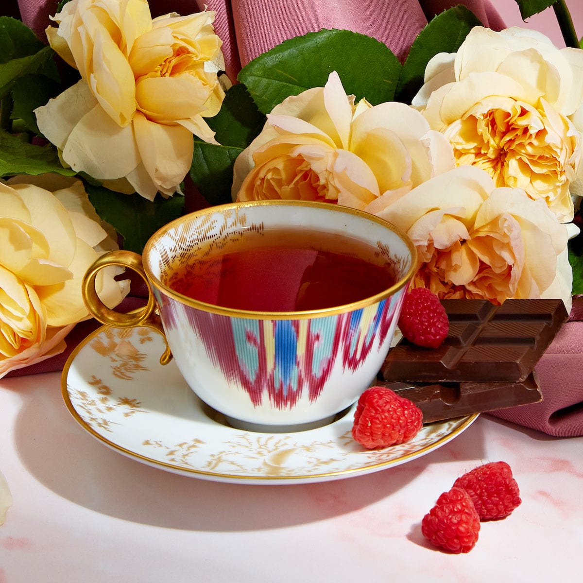 A porcelain teacup from the Dessert Lovers Starter Set, brimming with tea, rests on a matching saucer amid vibrant yellow roses and lush green leaves. Three raspberries and several pieces of dark chocolate, characteristic of the dessert sampler's delights, are artfully arranged beside the teacup and saucer on a pristine white surface.