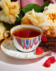 A porcelain teacup from the Dessert Lovers Starter Set, brimming with tea, rests on a matching saucer amid vibrant yellow roses and lush green leaves. Three raspberries and several pieces of dark chocolate, characteristic of the dessert sampler's delights, are artfully arranged beside the teacup and saucer on a pristine white surface.