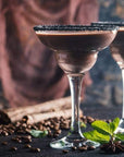 A close-up of two elegant cocktail glasses filled with a creamy chocolate beverage. The rims are coated with a dark substance, possibly sugar. Scattered coffee beans, cinnamon sticks, star anise, and mint leaves decorate the dark surface, evoking the enchanting aroma of Queen of the South: Delicious Cocoa Detox Tea by Magic Hour.