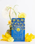 A vibrant blue packet labeled "Magic Hour: Queen of Ukraine: Spring Blossom Tea" with yellow and white floral designs stands against a white background. It is adorned with yellow and white flowers, including roses and anthuriums, enhancing the colorful and elegant presentation of this exquisite loose leaf tea.