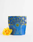 A blue tea packaging with gold floral patterns reads "Queen of Ukraine: Spring Blossom Tea, Magic Hour." The bag, adorned with a sun, moon, and stars design, sits beside a bright yellow flower on a plain white background. Experience the enchantment of Magic Hour's organic loose leaf blend.