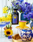 A vibrant arrangement of yellow and purple flowers surrounds an assortment of colorful teacups, a teapot filled with Queen of Ukraine: Spring Blossom Tea by Magic Hour, and a bottle with a decorative label featuring blue and gold designs. A sunflower and a blue cocktail are also present, all set on a reflective white surface.