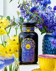 A vibrant arrangement of yellow and purple flowers surrounds an assortment of colorful teacups, a teapot filled with Queen of Ukraine: Spring Blossom Tea by Magic Hour, and a bottle with a decorative label featuring blue and gold designs. A sunflower and a blue cocktail are also present, all set on a reflective white surface.