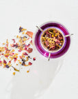 A top-view of a glass teacup filled with vibrant purple tea from the Luxe Tea Sampler Sips: The Queens Collection, accompanied by a metal tea infuser containing an assortment of caffeine-free herbal blends and healing botanicals. Loose dried herbs and flower petals from the collection are scattered on the white surface next to the teacup.