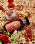 A box of Magic Hour's Queen of the Harvest: Herbal Relaxation Tea is surrounded by a glass teapot filled with organic tea, a honeycomb with a golden spoon, two traditional tea glasses brimming with loose leaf tea, red and white flowers, and greenery on a speckled countertop. The scene exudes a cozy and inviting atmosphere.