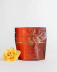 A pouch of **Queen of the Harvest : Herbal Relaxation Tea** labeled **Magic Hour** with an intricate gold design stands against a white background. To the left of the pouch, there is a vibrant yellow flower placed for decoration, enhancing the allure of this organic loose leaf tea.