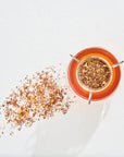 Overhead view of a cup of Luxe Tea Sampler Sips: The Queens Collection from Magic Hour with loose leaf tea in a strainer. Some of the organic tea leaves are scattered on a white surface beside the cup. The tea has an orange hue.