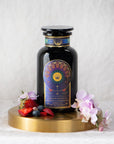 A dark glass jar labeled "Queen of the Rainforest : Cacao-Berry Tea" by Magic Hour sits on a circular wooden stand, surrounded by fresh strawberries, blueberries, and delicate purple and white flowers. The organic tea promises a delightful experience. In the background is a soft white cloth, enhancing the serene ambiance.