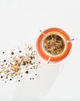 Top-down view of a glass containing brown-colored organic tea, with a stainless steel infuser filled with loose leaf tea, placed on a white surface. Loose tea leaves and berries are scattered around the glass. The product featured is Luxe Tea Sampler Sips: The Queens Collection by Magic Hour.