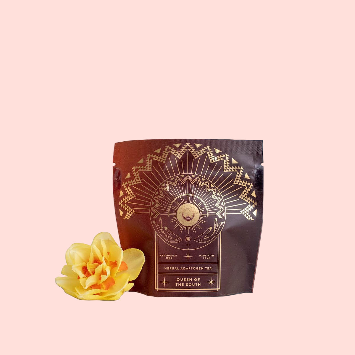 A sealed brown pouch labeled "Queen of the South: Delicious Cocoa Detox Tea - Magic Hour" with decorative gold designs, featuring a sun, crescent moon, and geometric patterns. Beside the Magic Hour pouch on a white surface lies a vibrant yellow flower.