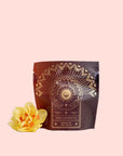 A sealed brown pouch labeled "Queen of the South: Delicious Cocoa Detox Tea - Magic Hour" with decorative gold designs, featuring a sun, crescent moon, and geometric patterns. Beside the Magic Hour pouch on a white surface lies a vibrant yellow flower.