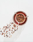 A clear glass teacup filled with a dark reddish liquid sits on a white surface. The teacup contains a tea infuser with Magic Hour's Luxe Tea Sampler Sips: The Queens Collection, a mix of dried leaves and spices. Some of the leaves and spices are scattered beside the cup.