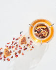 A top view of a glass vessel containing Luxe Tea Sampler Sips: The Queens Collection herbal tea with dried citrus slices and floral petals. Some of the healing botanicals are scattered artistically to the left of the vessel on a white surface, casting soft shadows.