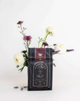 A dark-colored packet labeled "Club Magic Hour: Raspberry Earl White Tea" is placed against a white background, partially surrounded by a small arrangement of white and purple flowers. The packet, filled with organic loose leaf tea, displays an intricate design featuring celestial imagery.