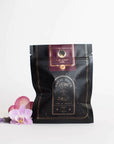 A black bag of Club Magic Hour Raspberry Earl White Tea with gold and red accents is displayed against a white background. The bag features elegant branding, including a logo and text. Bright purple and pink flowers are placed beside the bag for decoration, highlighting the loose leaf tea inside.
