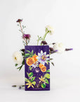 A beautifully designed coffee bag in a striking purple hue, adorned with vibrant illustrations of fruits and flowers, including a blend of passion fruit, sits gracefully in front of an exquisite floral arrangement featuring white roses, lavender, and other blossoms against a plain white background. This stunning display evokes the charm of mornings on the Amalfi Coast and perfectly showcases the Romance in Ravello - Chocolate-Passionfruit-Rose-Cream White Tea by Magic Hour.