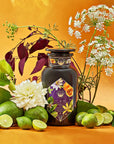 An elegant dark glass bottle labeled "Romance in Ravello" from Magic Hour is surrounded by fresh green limes, lime wedges, and assorted flowers against a vibrant yellow-orange background. This visually rich composition evokes the natural beauty and freshness of the Amalfi Coast, enhanced by hints of chocolate, passion fruit, and rose cream.